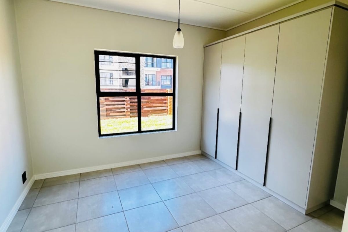 A bright, empty bedroom featuring neutral tiled flooring, a large built-in wardrobe, and a window with security bars.