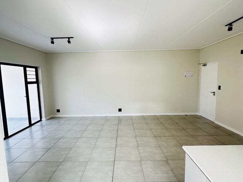 A spacious, empty open-plan living area featuring neutral tiled flooring, modern track lighting, and a sliding glass door leading to an outdoor space.
