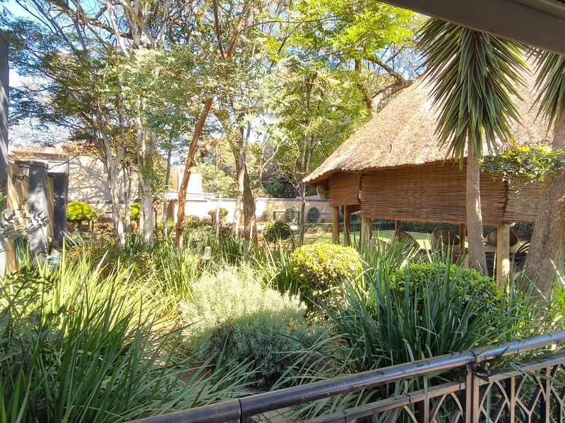 A lush, established garden featuring a traditional thatched lapa, mature trees, and a wrought iron balcony railing overlooking the outdoor space.