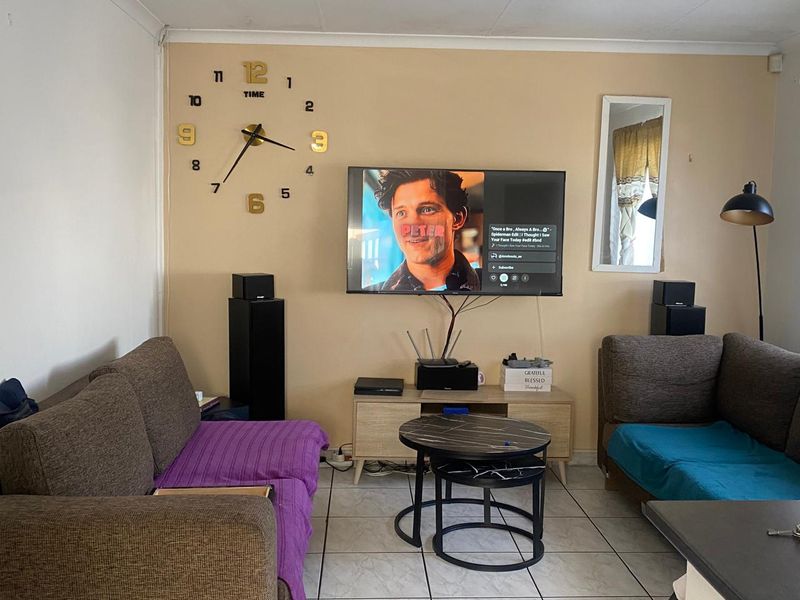 A living room featuring tiled flooring and a wall-mounted television. The space is functional but would benefit from cosmetic updates and cable management.