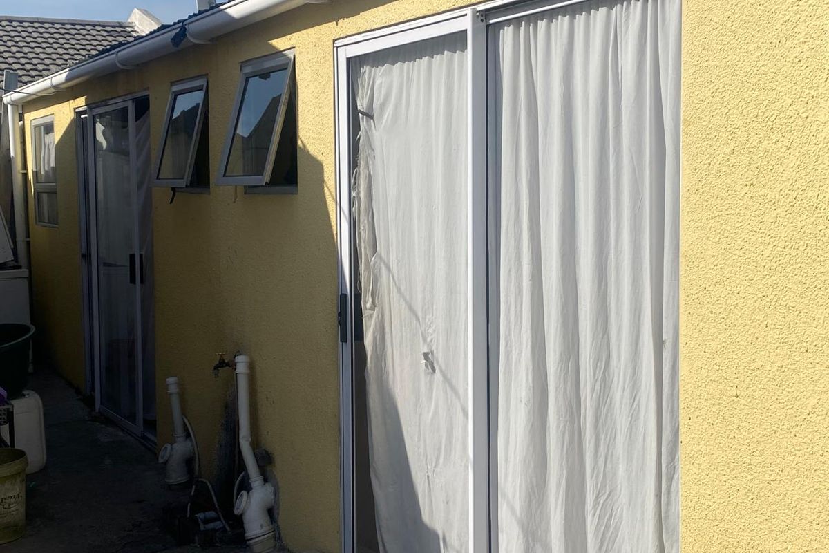 Rear exterior view of the property featuring sliding glass doors, top-hung windows, and exposed external plumbing fixtures along the yellow rendered wall.