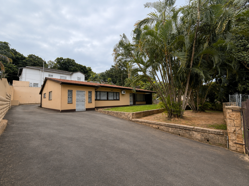 Front view of a single-story house featuring an asphalt driveway, tiled roof, and established garden with a retaining wall.