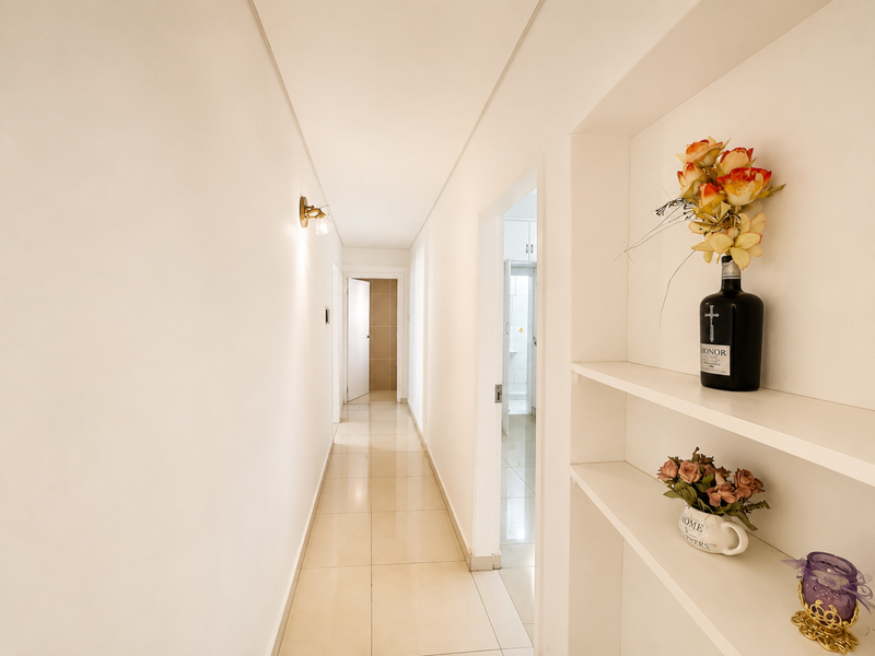 A bright, clean hallway featuring neutral tiled flooring, built-in wall shelving, and modern wall-mounted lighting, providing access to adjacent rooms.