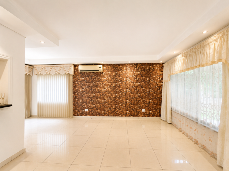 Spacious living area featuring tiled flooring, a decorative brick-effect feature wall, recessed ceiling lighting, and a wall-mounted air conditioning unit.