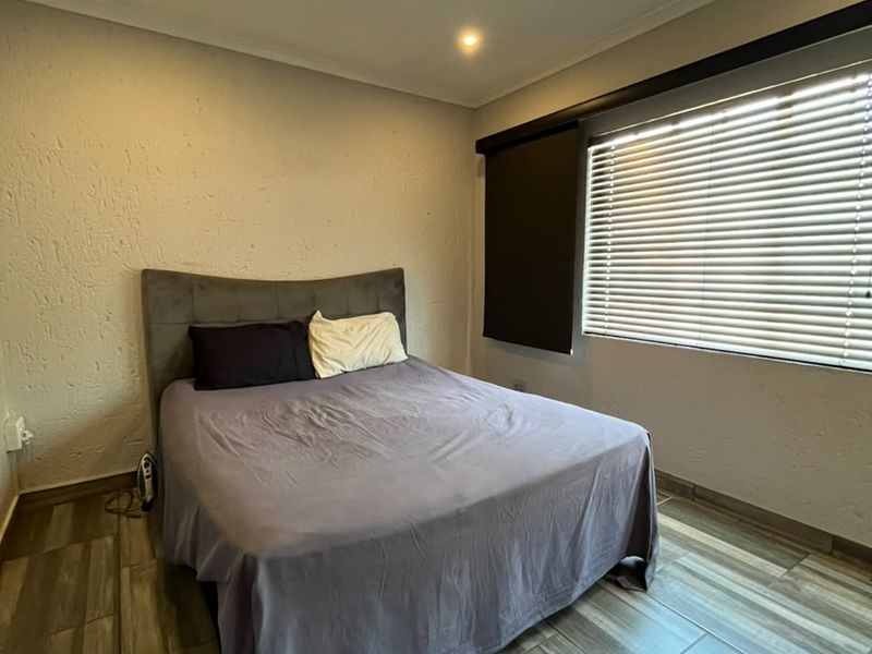 A bedroom featuring modern wood-look tiled flooring, recessed downlighting, and venetian blinds, providing a clean and functional living space.