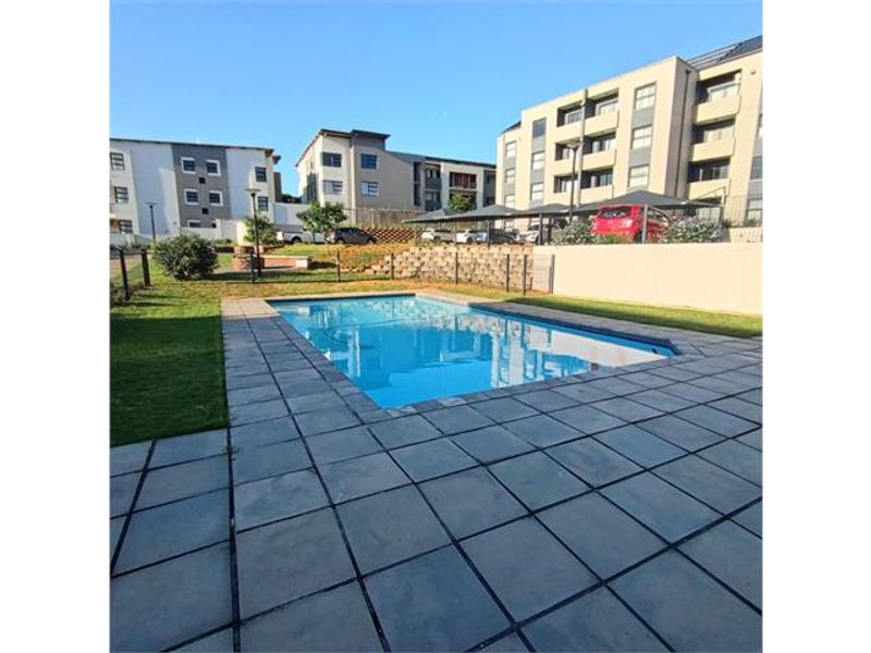 A well-maintained communal swimming pool area featuring a paved deck, retaining wall, and surrounding lawn within an apartment complex.