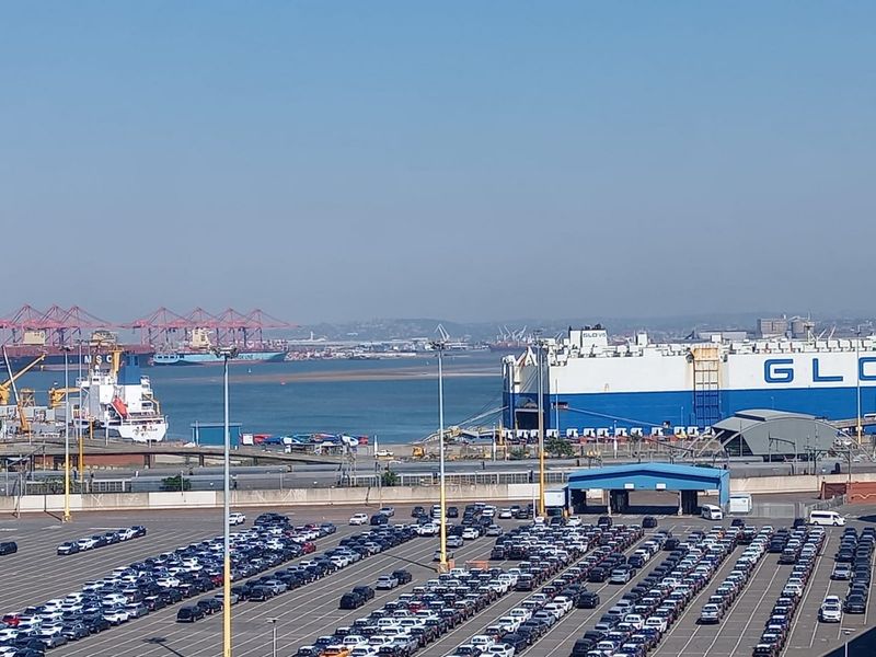 An elevated view overlooking a busy industrial harbor with large cargo ships and a vehicle storage terminal.