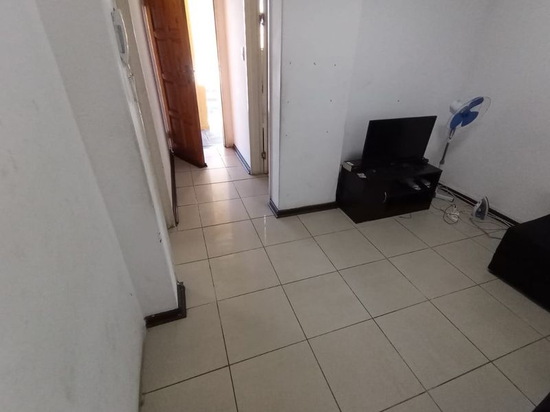 A compact living area featuring neutral tiled flooring and an intercom system, showing signs of wear on the walls and skirting boards.