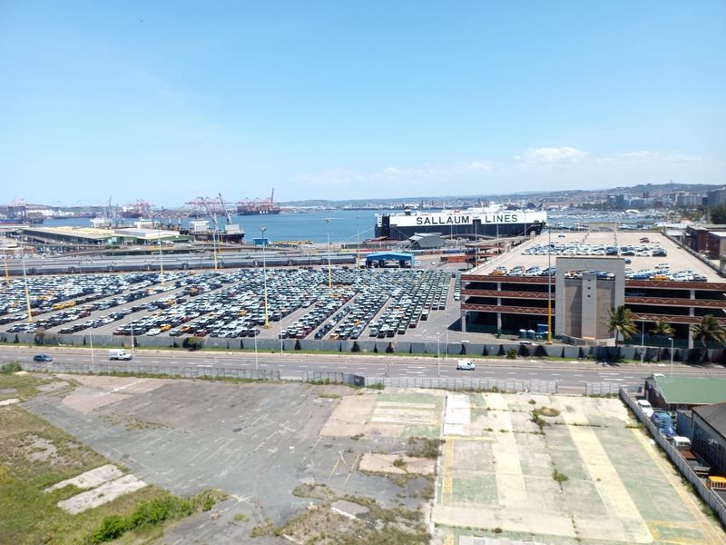An elevated view overlooking a large industrial port area, vehicle storage facility, and coastal harbor infrastructure.