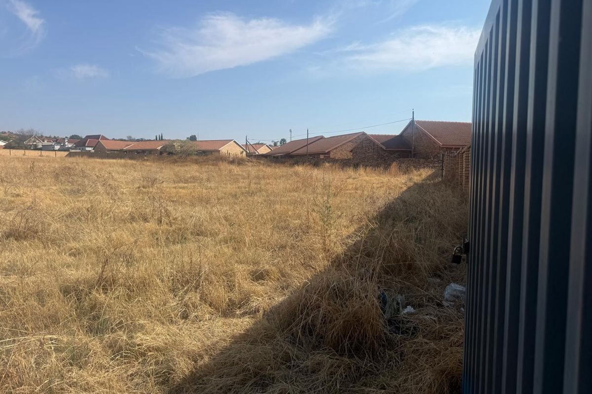 A vacant residential plot featuring dry, overgrown grass and perimeter fencing, offering a clear view of the surrounding neighborhood.