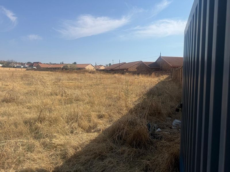 A vacant residential plot featuring dry, overgrown grass and perimeter fencing, offering a clear view of the surrounding neighborhood.
