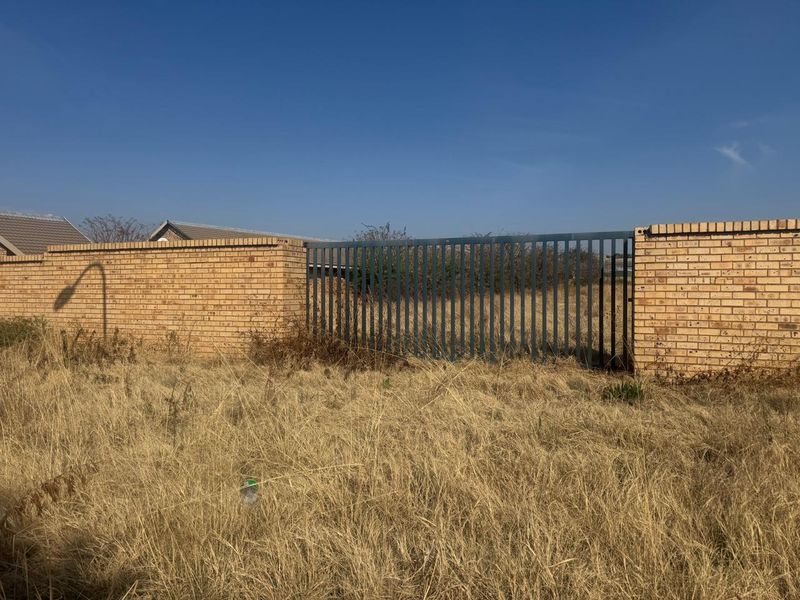 A view of a vacant residential plot featuring a brick boundary wall and a metal gate, surrounded by dry, overgrown grass.