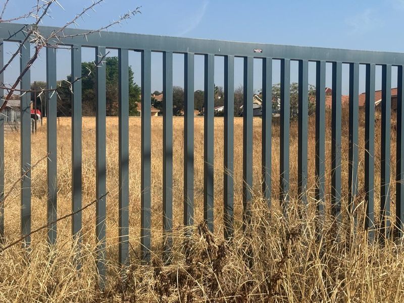A view of a vacant residential plot secured by metal perimeter fencing, showing dry grass and surrounding neighborhood houses.