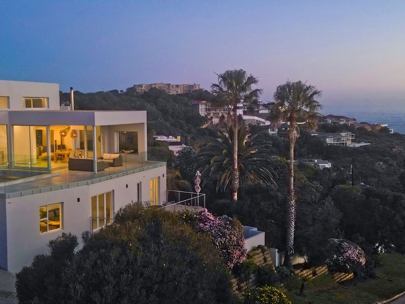 Aerial view of a modern multi-level residence featuring expansive terraces, glass balustrades, and scenic coastal hillside surroundings at dusk.