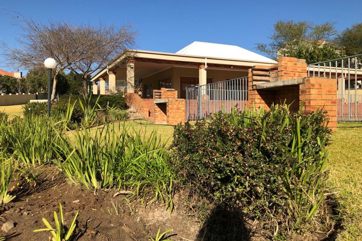 Rear view of the property featuring a covered patio, brick built-in braai area, and established garden with metal security fencing.