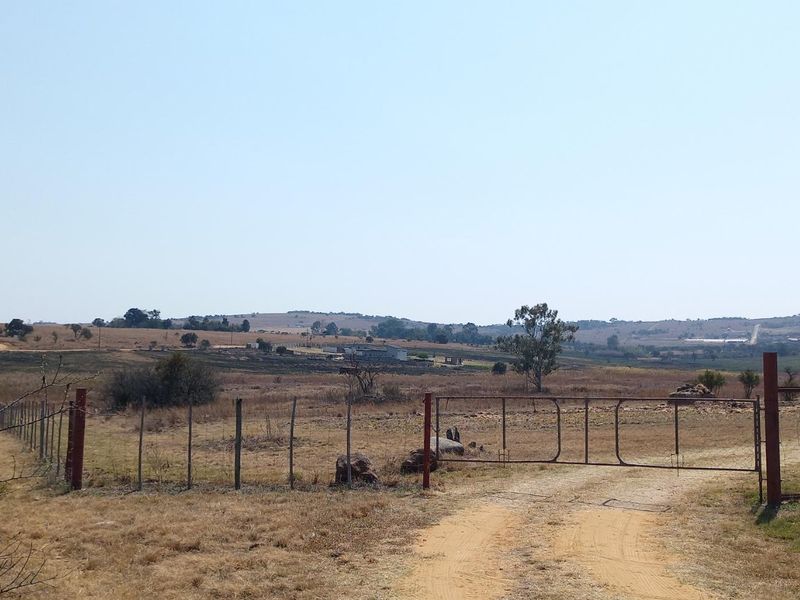 A view of a vacant residential plot featuring wire fencing, a metal gate, and a dirt access road leading into the open landscape.