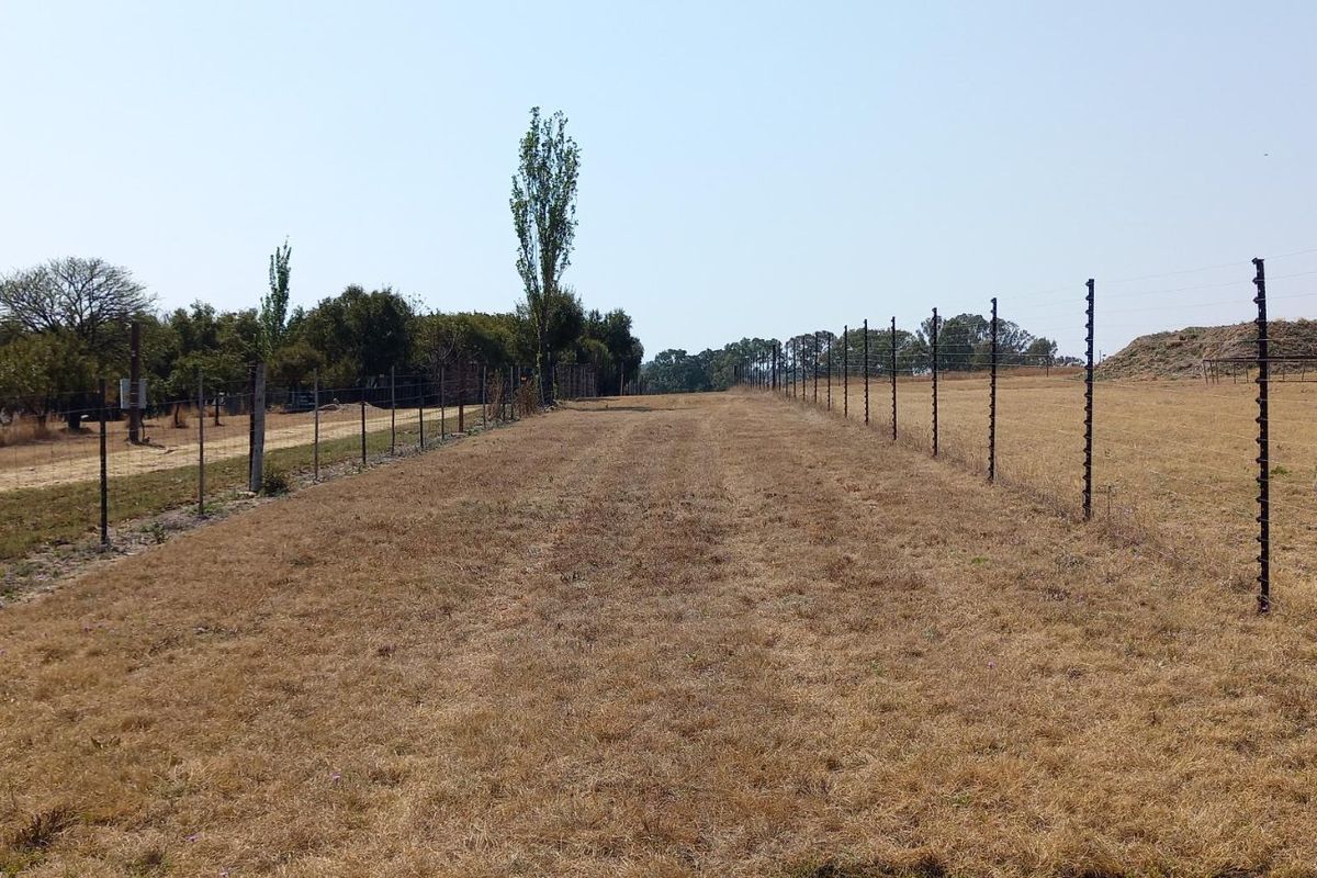 A view of a vacant residential plot featuring dry grass, perimeter electric fencing, and a water storage tank.