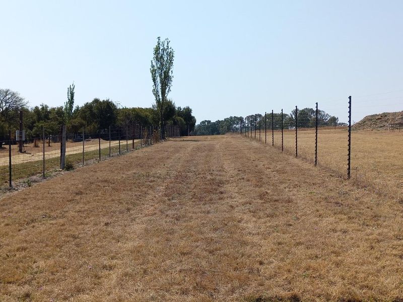 A view of a vacant residential plot featuring dry grass, perimeter electric fencing, and a water storage tank.