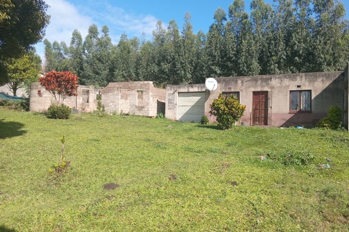 Rear view of a property featuring an existing dwelling with a garage and an adjacent unfinished masonry structure set on a large grassed plot.