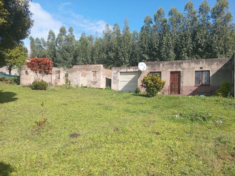 Rear view of a property featuring an existing dwelling with a garage and an adjacent unfinished masonry structure set on a large grassed plot.