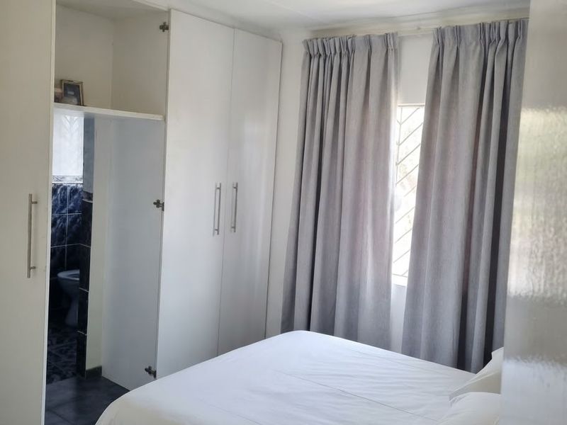 A bedroom featuring built-in cupboards and a window with security bars, providing direct access to an en-suite bathroom area.