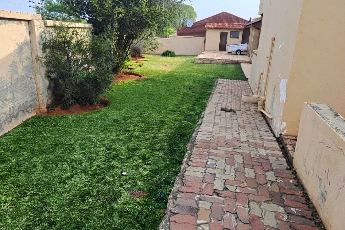 A rear garden area featuring a paved walkway, lawn, and boundary walls, showing signs of wear and maintenance requirements on the exterior walls.