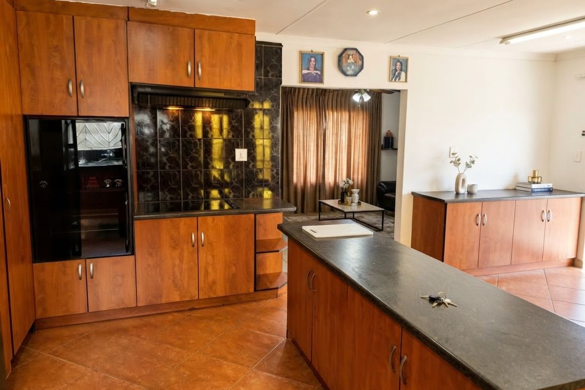 A functional kitchen featuring wood-finish cabinetry, dark laminate countertops, a built-in hob with extractor, and tiled flooring, opening into an adjacent living area.