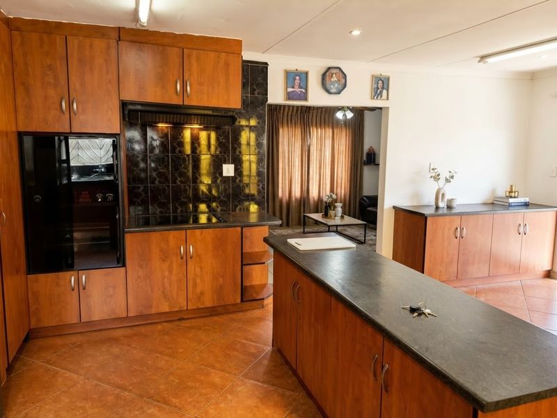 A functional kitchen featuring wood-finish cabinetry, dark laminate countertops, a built-in hob with extractor, and tiled flooring, opening into an adjacent living area.