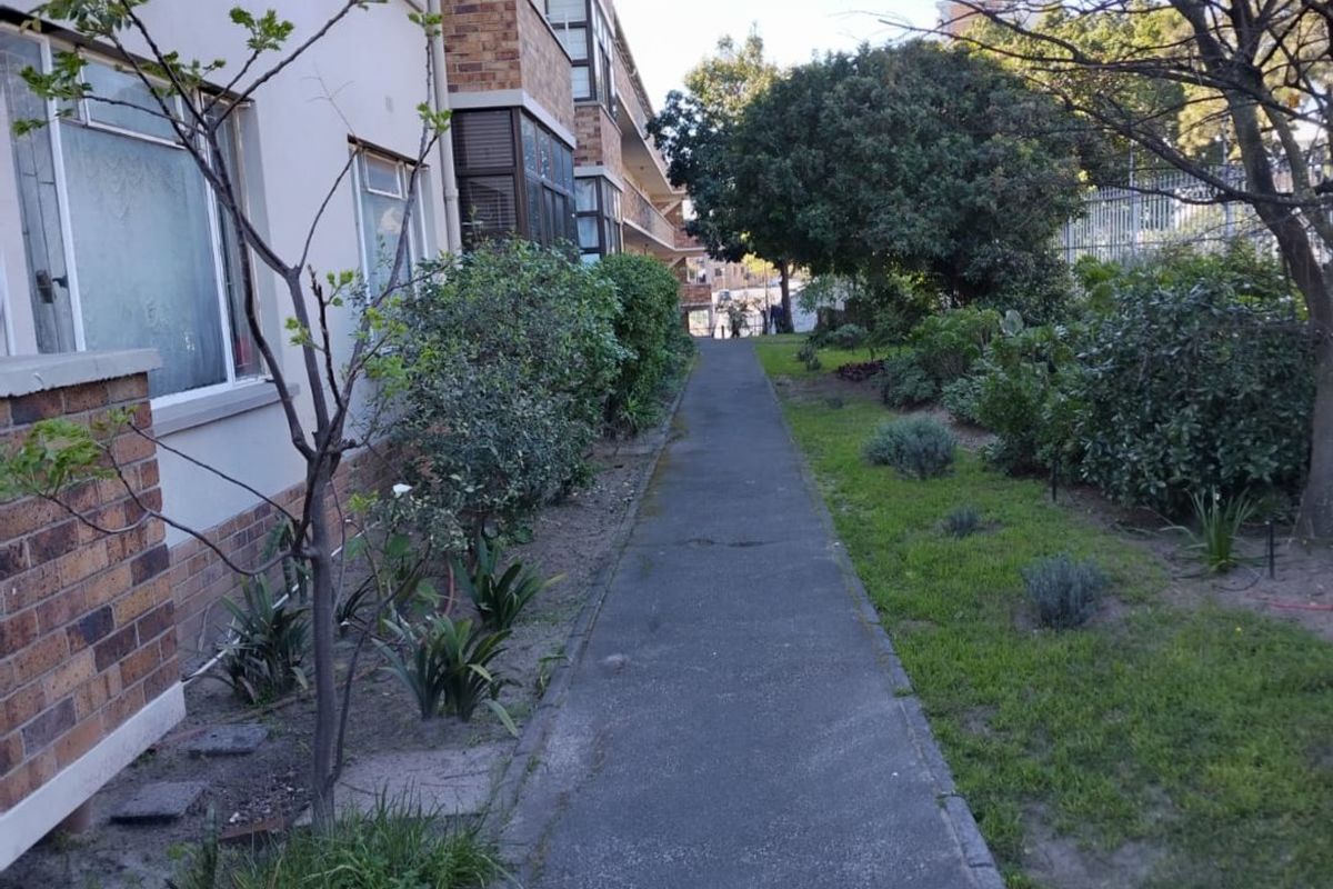 A view of the apartment building's rear exterior, featuring a concrete walkway alongside a landscaped garden and brick facade.