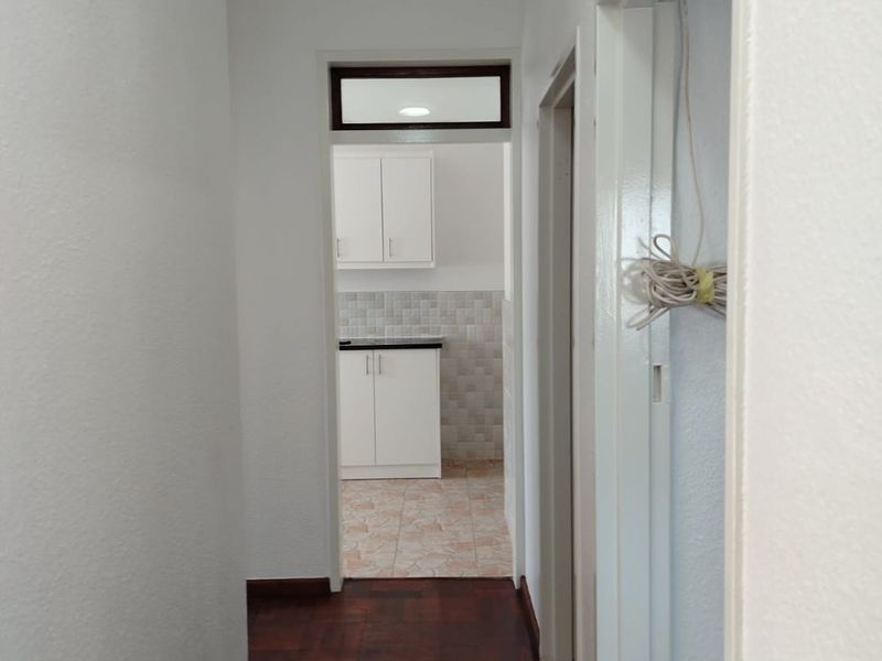 A clean hallway with dark parquet flooring leading into a kitchen area featuring white cabinetry and tiled splashbacks.