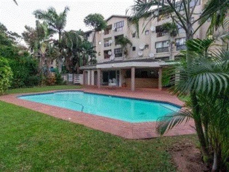 A communal swimming pool area featuring a paved deck, covered patio space, and surrounding landscaped gardens within an apartment complex.