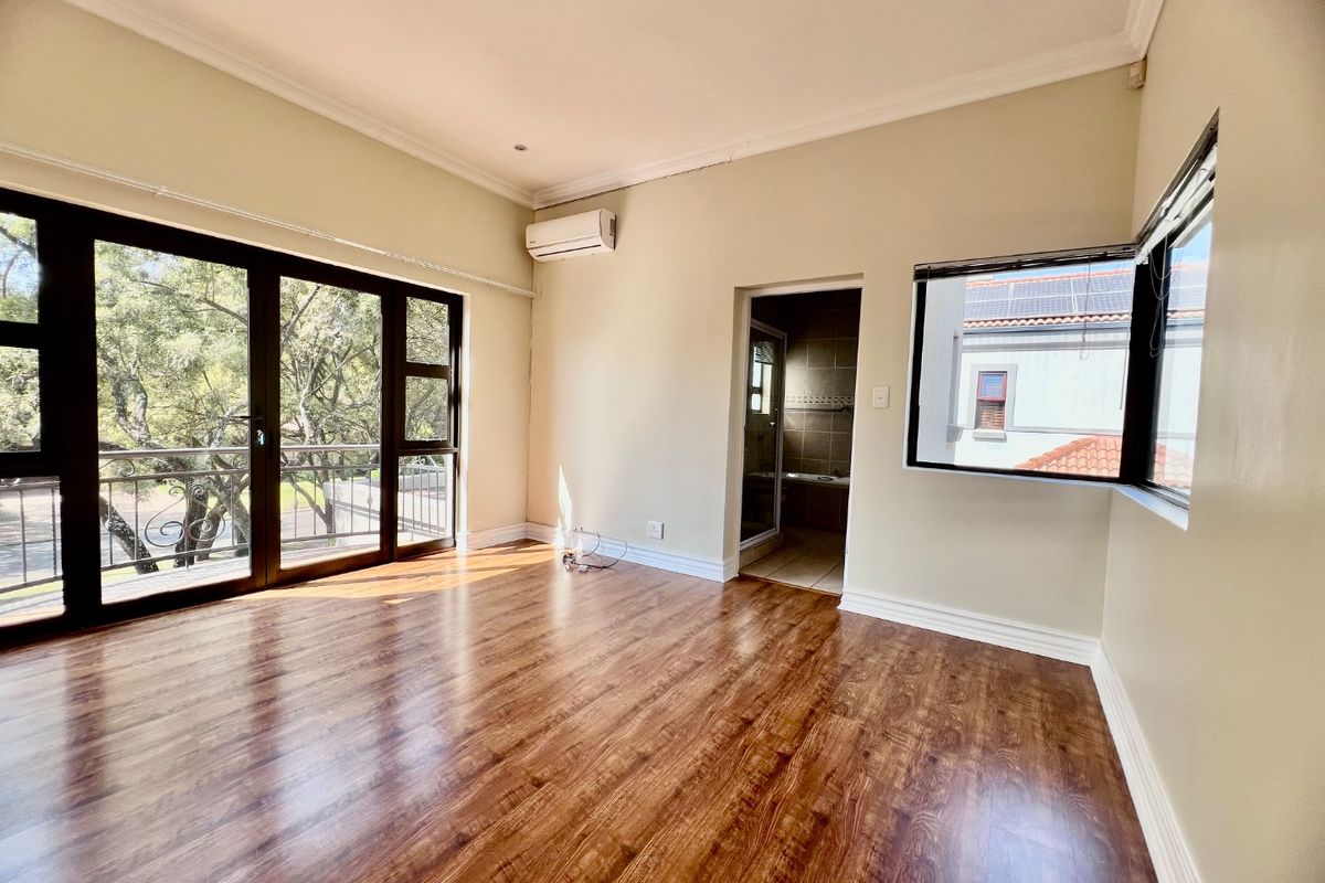 A bright, empty bedroom featuring laminate flooring, a split-system air conditioner, and sliding glass doors leading to a balcony, with an adjoining en-suite bathroom.