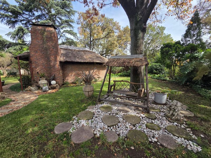 A lush, established garden featuring a rustic brick structure with a thatched roof, mature trees, and a stone-paved pathway.