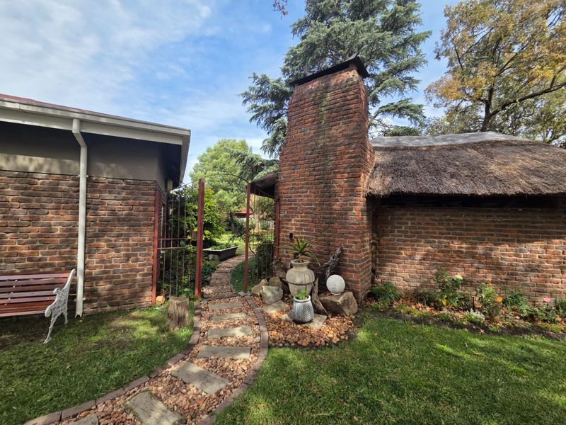 A charming rear garden area featuring a rustic brick chimney, a thatched roof structure, and a stone pathway leading through a wrought iron gate.