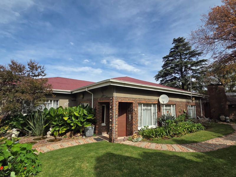 Front elevation of a single-story brick house featuring a red metal roof, established garden, and paved pathway leading to the entrance.