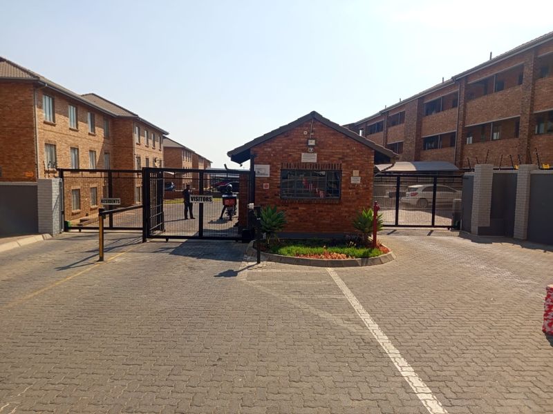 The apartment complex entrance features a secure gated access point with a brick guard house, paved driveway, and perimeter electric fencing.