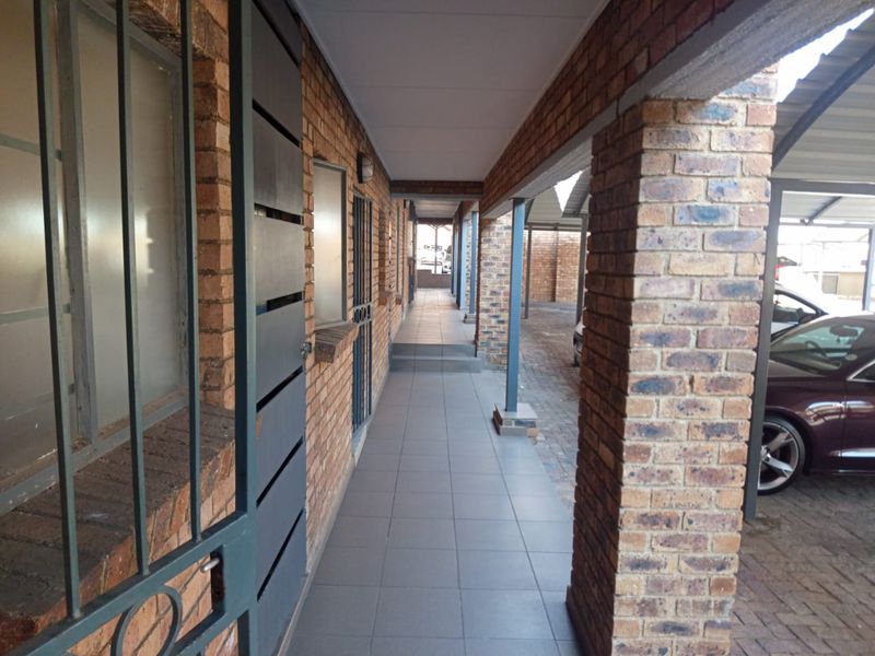 A covered exterior walkway featuring brick construction and tiled flooring, providing access to the apartment entrance and adjacent parking area.