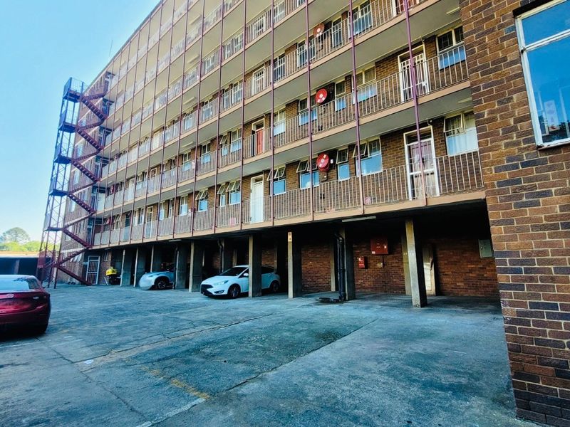 Exterior view of a multi-story apartment building featuring brick facade, metal balcony railings, an external fire escape, and a ground-level covered parking area.