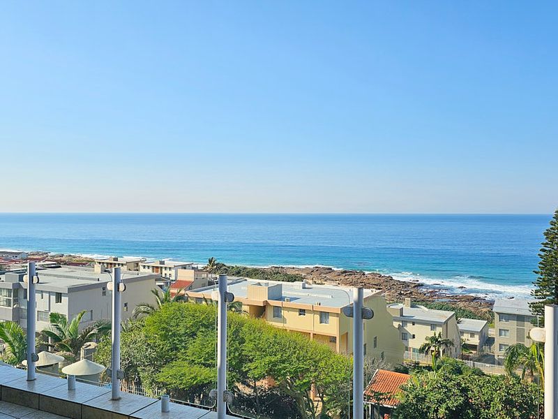 A clear, elevated coastal view overlooking the ocean, surrounding residential buildings, and lush greenery from a private balcony with a glass balustrade.