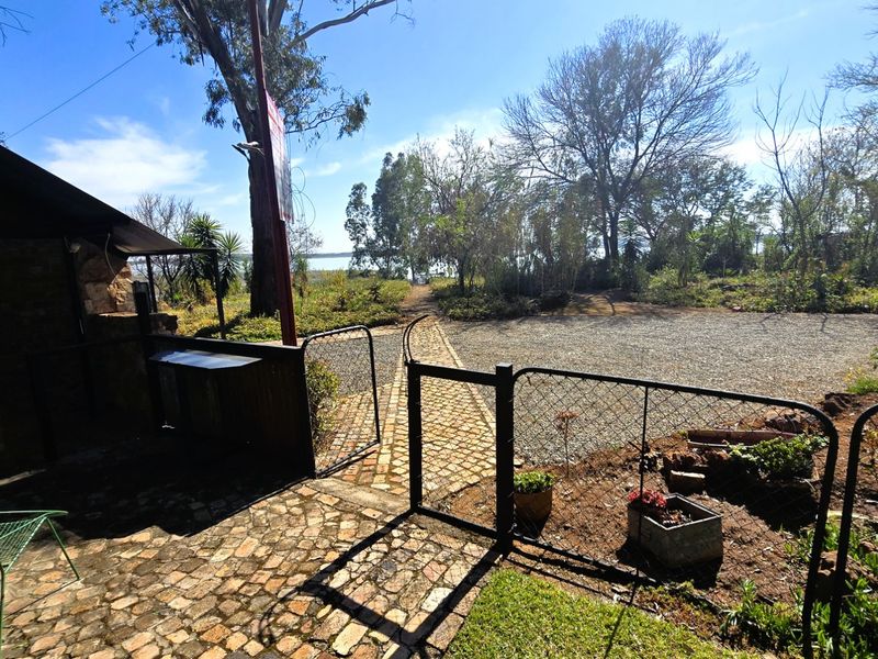 Rear outdoor area featuring a brick-paved patio with a built-in braai, gravel driveway, and fenced garden space with mature trees.