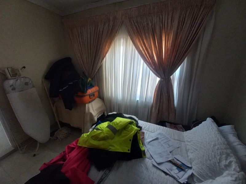 A bedroom featuring tiled flooring and window curtains, currently cluttered with personal items.