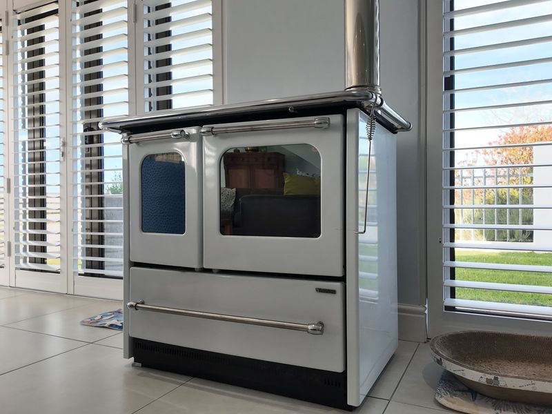 A bright living area featuring a classic white freestanding wood-burning stove, tiled floors, and large glass doors with white plantation shutters.