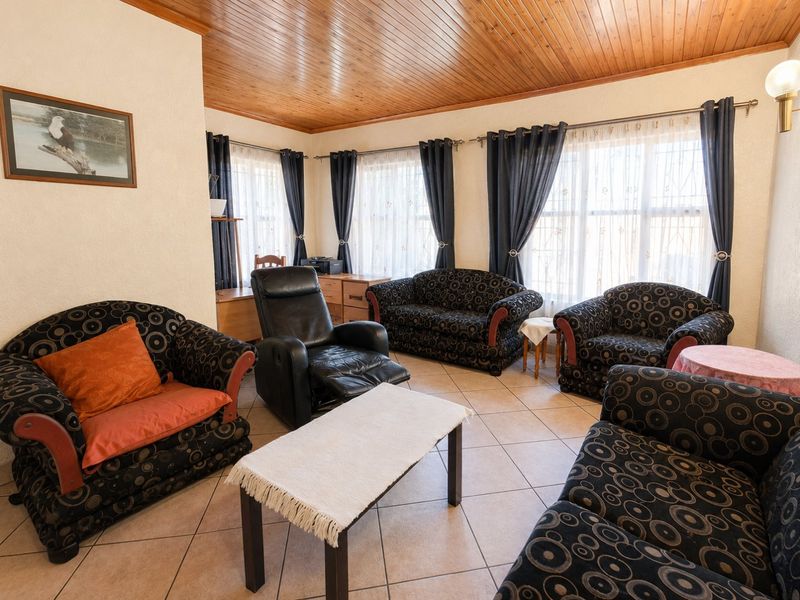 A spacious living room featuring tiled flooring and a warm timber tongue and groove ceiling, illuminated by natural light through large windows.