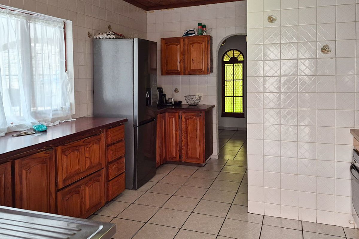 A dated kitchen featuring wooden cabinetry, tiled walls and flooring, a built-in oven, and a hob, leading through an arched doorway.