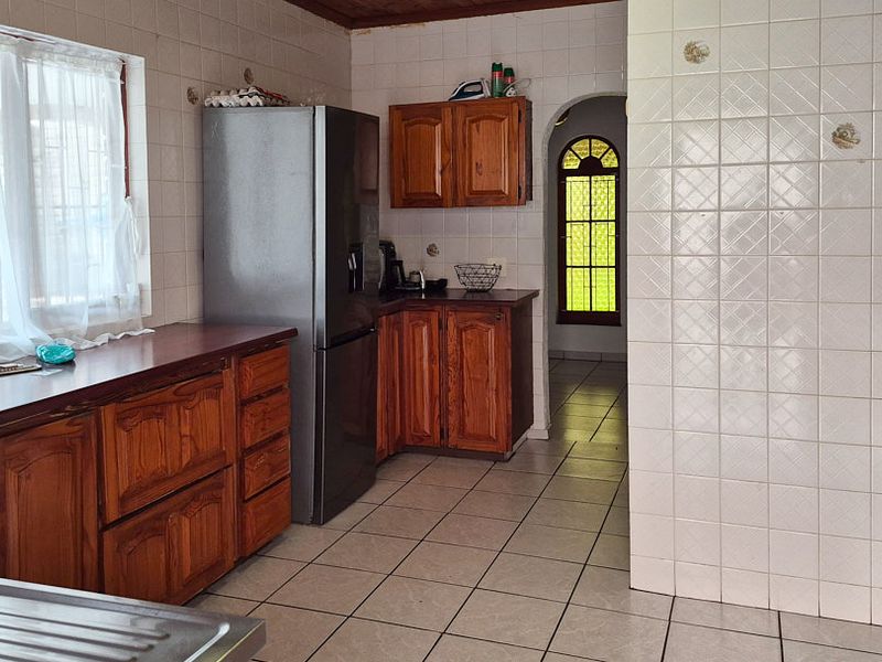 A dated kitchen featuring wooden cabinetry, tiled walls and flooring, a built-in oven, and a hob, leading through an arched doorway.