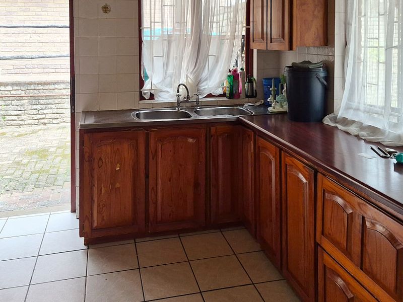 A functional kitchen featuring wooden cabinetry, a double stainless steel sink, tiled flooring, and direct access to the exterior through a security gate.