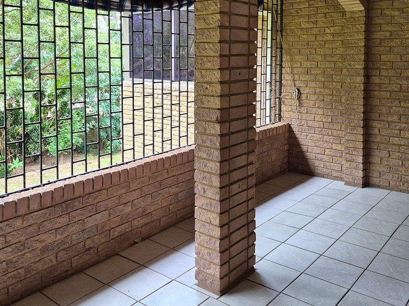 A covered patio area featuring face brick walls, tiled flooring, and security bars, offering a view of the surrounding garden.