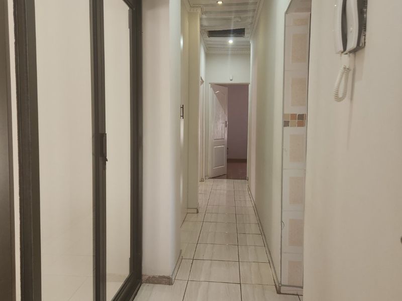 A long, tiled hallway featuring recessed lighting, an intercom system, and a sliding glass door, leading to an adjacent room.