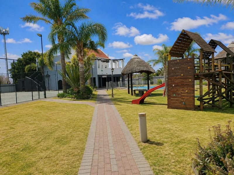 Communal estate facilities featuring a paved walkway, tennis court, playground, thatched gazebo, and swimming pool set within a well-maintained landscaped garden.