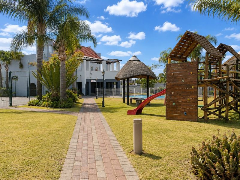 A well-maintained communal garden area featuring a paved walkway, thatched gazebo, and playground equipment, set within a secure townhouse complex.